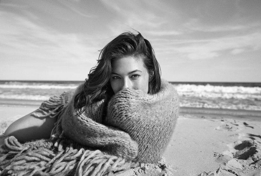 A young woman with wavy hair covered with a thick knit blanket at the beach, black and white photo, capturing natural beauty and relaxed seaside atmosphere.