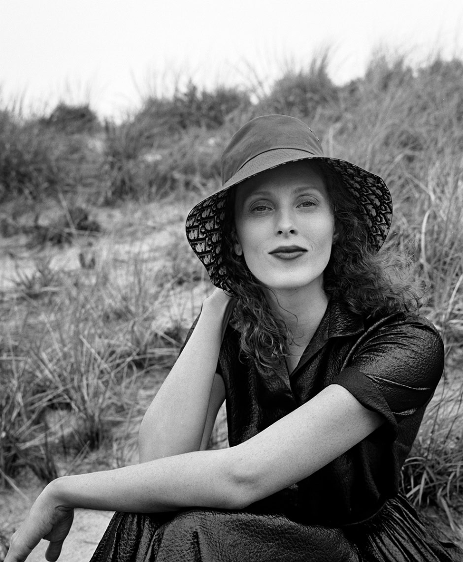 Black and white portrait of a woman wearing a wide-brimmed hat and vintage style dress sitting outdoors on a grassy dune, fashion photography, natural light, elegant and timeless style, Pamela Hanson Photography.