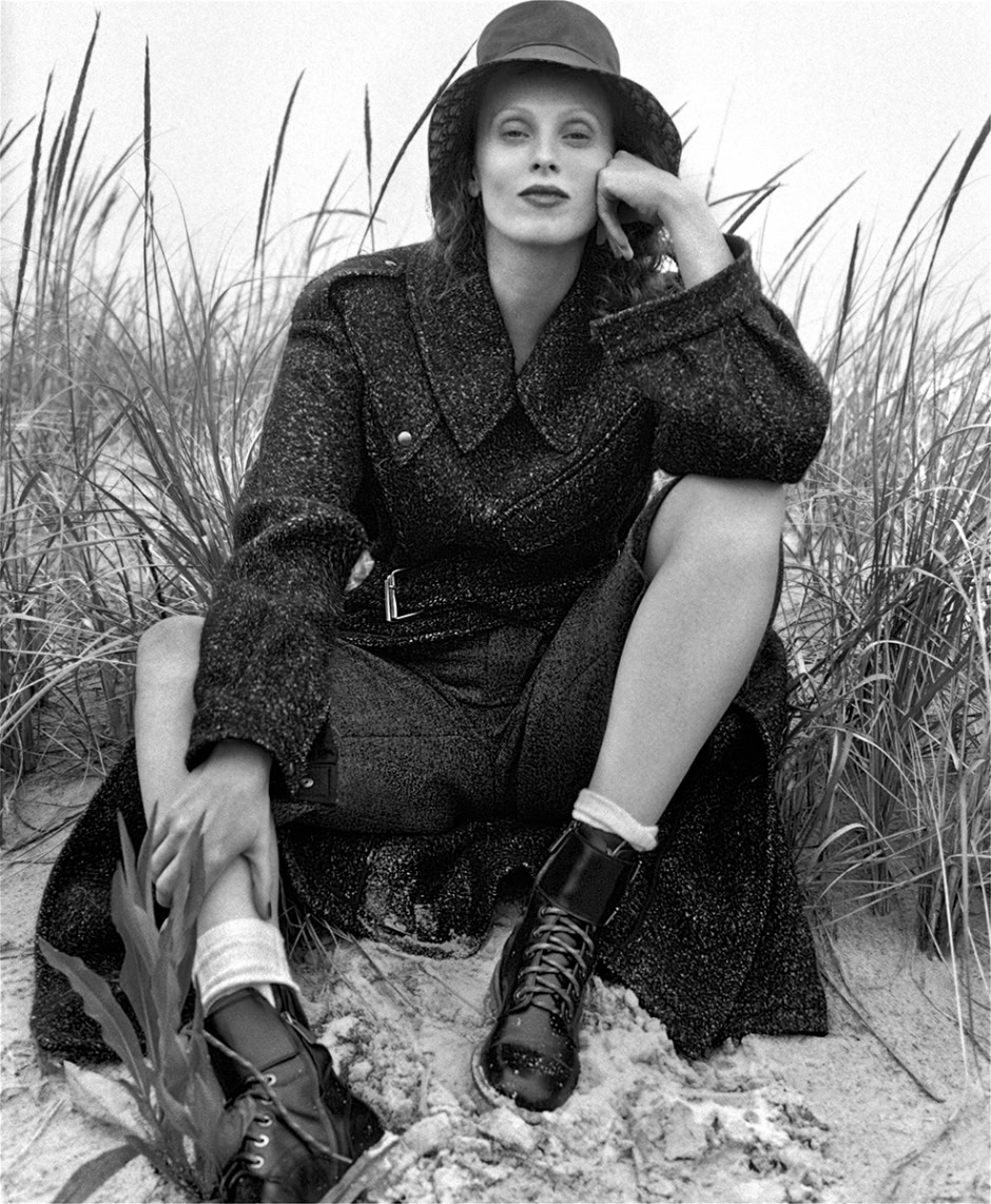 Black and white fashion photography of a woman sitting in sand dunes with tall grass, styled in a textured coat and hiking boots, capturing elegant outdoor portrait by Pamela Hanson.