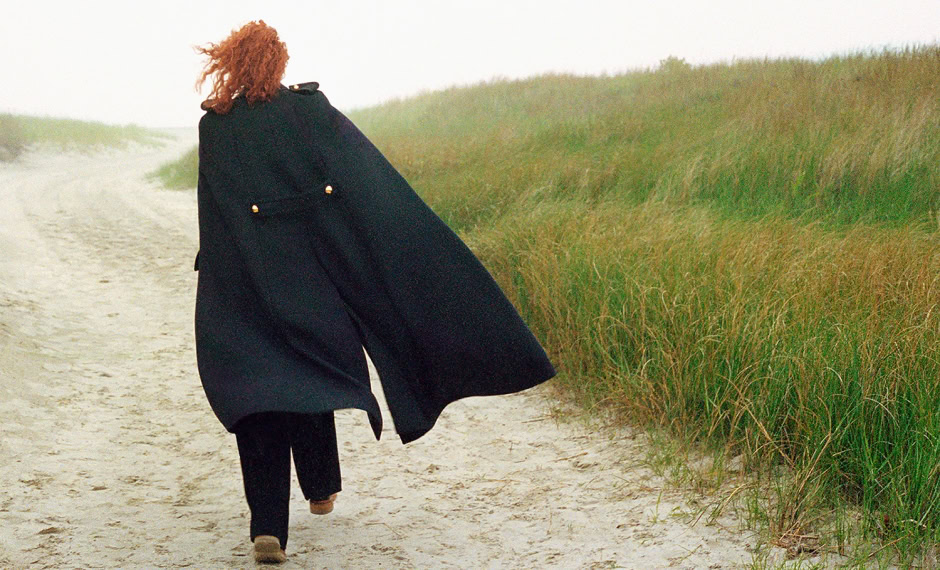 Woman walking along a sandy beach with green dunes in the background, wearing a black coat and pants, captured by Pamela Hanson Photography for stylish fashion and lifestyle photography, environmental portraiture.