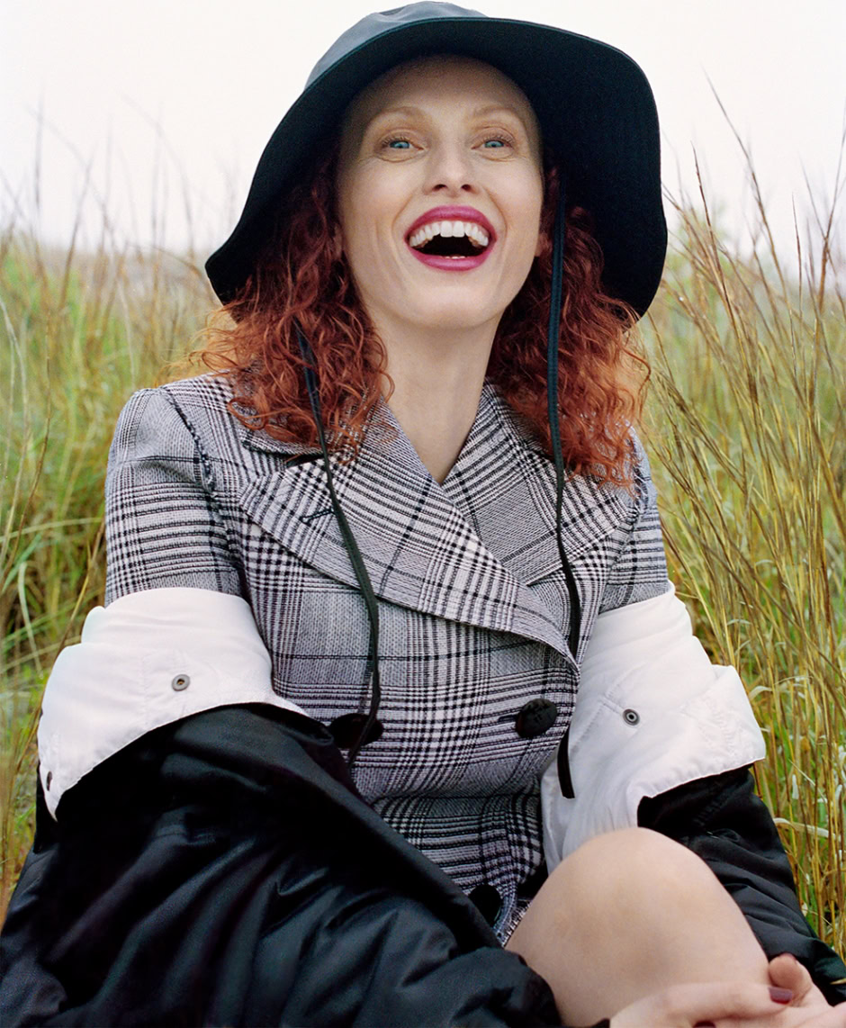Portrait of a smiling woman wearing a wide-brimmed hat and plaid coat, sitting in a grassy field, high-quality fashion photography, natural light, outdoor portrait, Pamela Hanson Photography.