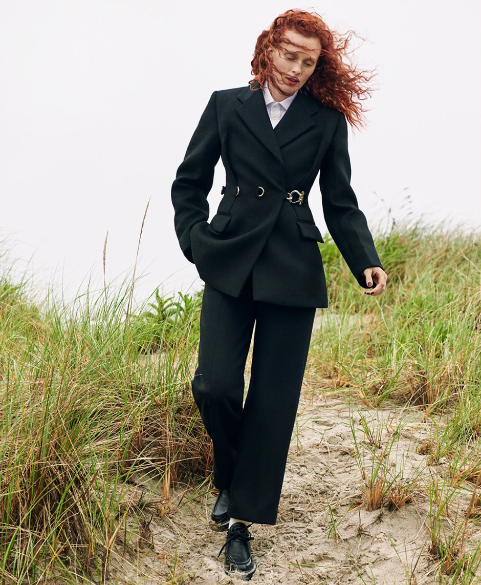 Elegant woman in tailored black suit walking on a sandy beach with green grasses, showcasing professional fashion photography.