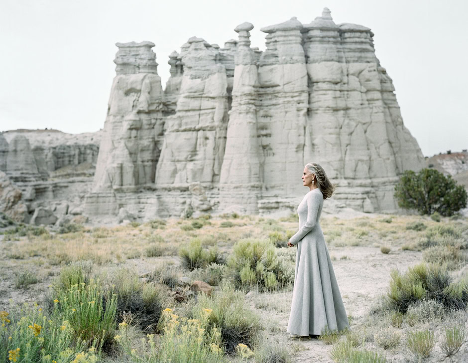 Elderly woman in a long, elegant grey dress standing amidst desert vegetation with towering rock formations behind her, capturing a moment of serenity and natural beauty.