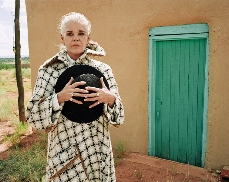 Elegant woman in black and white checkered coat holding a wide-brimmed hat, outdoors with a rustic turquoise door and natural landscape, showcasing fine art fashion photography.