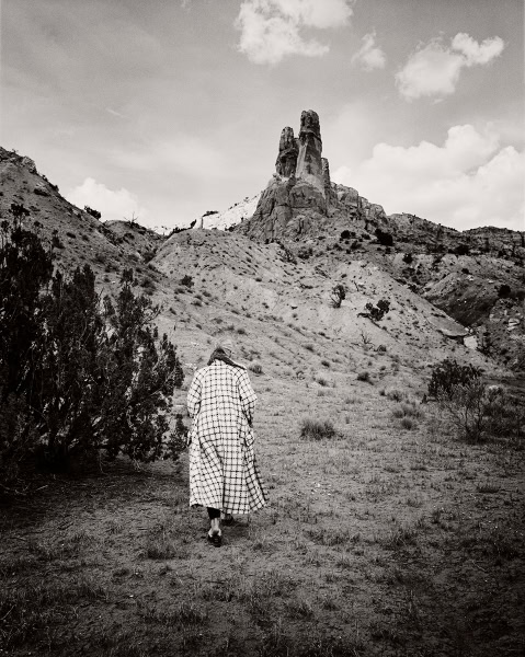 A black and white landscape photograph of a woman walking towards a rugged mountain formation with dramatic rock spires, taken by Pamela Hanson Photography.