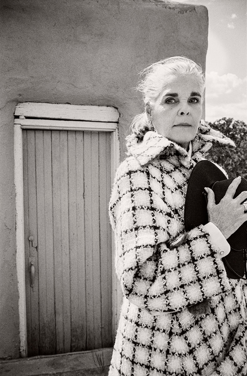 Elegant black and white portrait of an older woman with silver hair, wearing a stylish checkered coat, standing outdoors near a rustic door, showcasing timeless fashion photography.