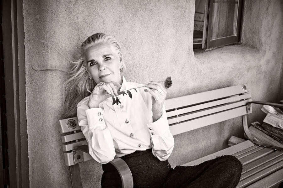 Elegant black-and-white portrait of a mature woman with flowing hair, sitting on a park bench with a contemplative expression, holding a flower, showcasing timeless beauty and sophisticated style.