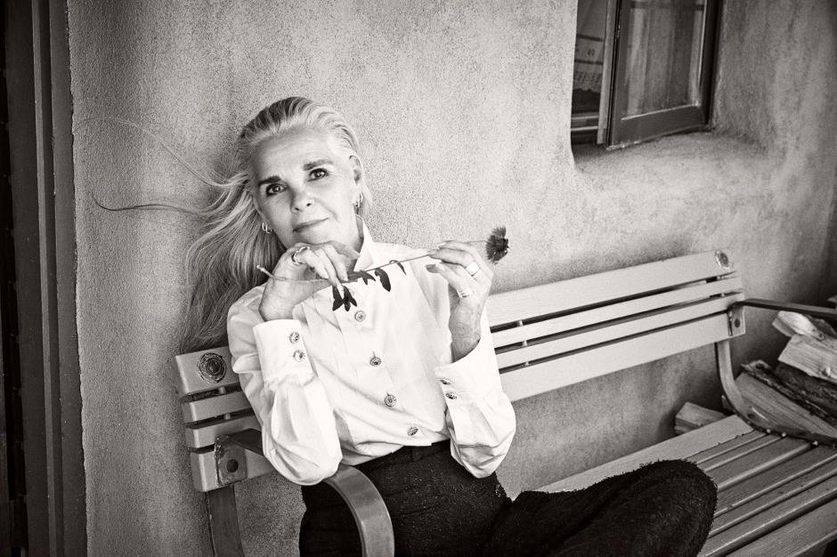 Elegant black-and-white portrait of a mature woman with flowing hair, sitting on a park bench with a contemplative expression, holding a flower, showcasing timeless beauty and sophisticated style.