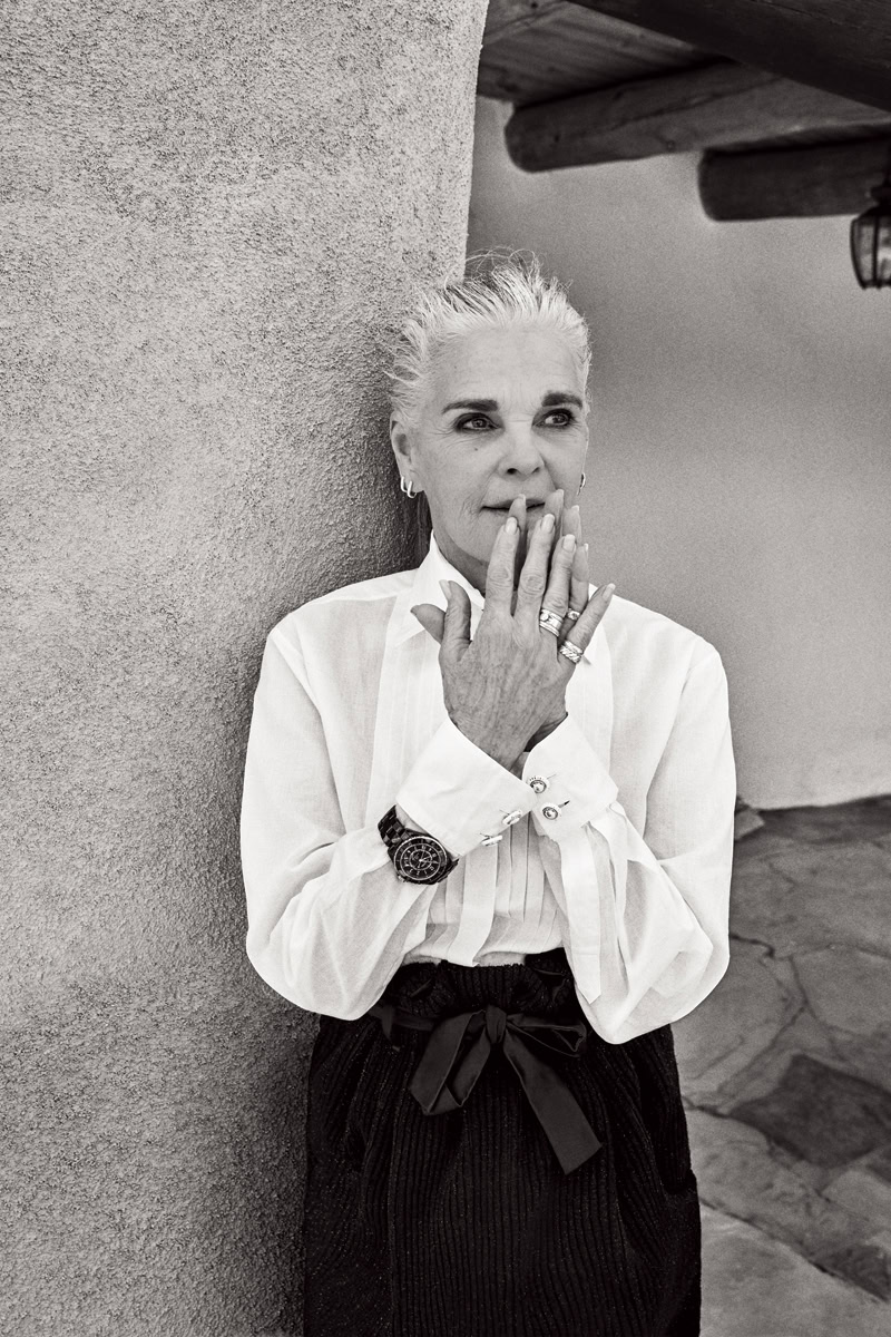 Elegant elderly woman with short white hair in a stylish white blouse and black trousers, standing against a textured wall, showcasing fashion, confidence, and timeless style.