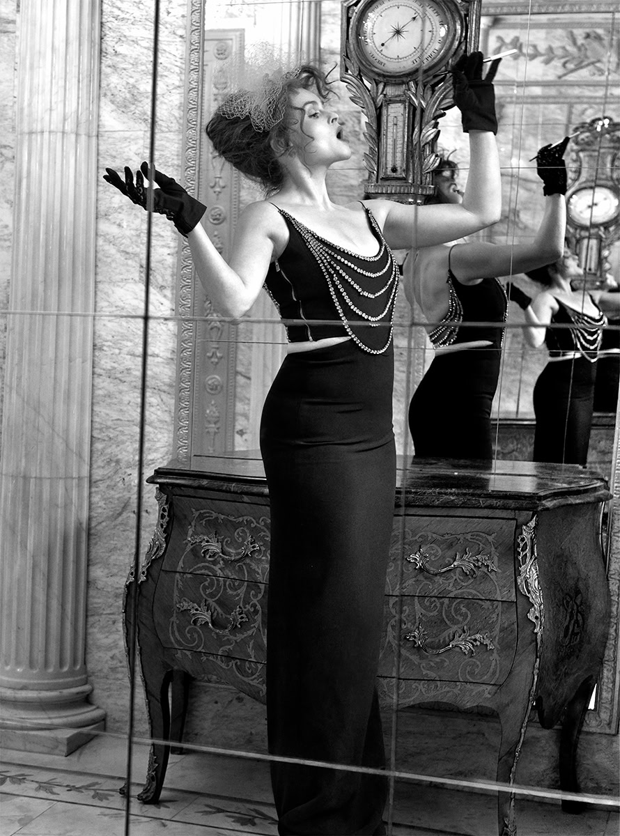 Elegant woman in vintage black evening gown with pearl embellishments, standing by an ornate antique dresser, reflected in a mirror, showcasing high fashion and classic Hollywood glamour photography.