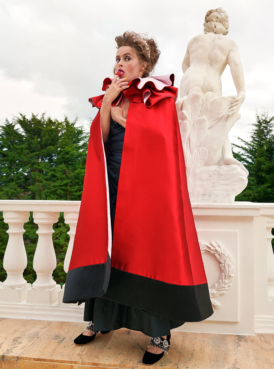 Elegant woman in red and black evening gown with ruffled collar, standing outdoors near white classical statue, stylish fashion photography by Pamela Hanson for artistic and commercial portfolio.