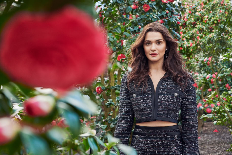 Vibrant portrait of a woman in a stylish, designer tweed outfit standing among blooming camellia bushes, capturing elegance, fashion photography, and natural beauty.
