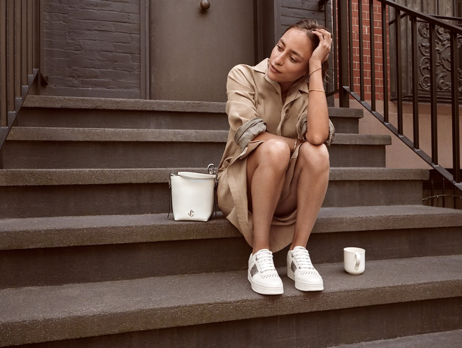 Relaxed woman sitting on brownstone steps with white sneakers, a beige trench coat, a white handbag, and a coffee mug, captured by Pamela Hanson Photography in urban street style.