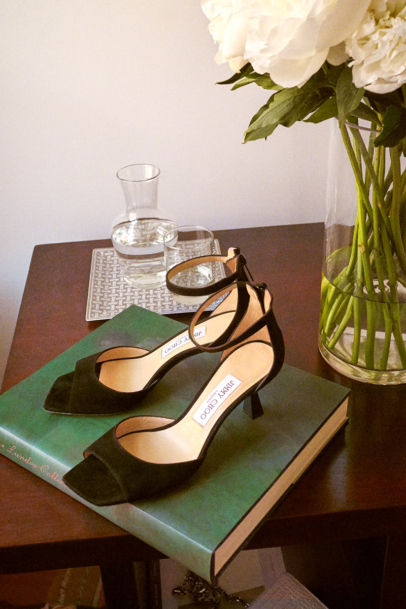Elegant black Jimmy Choo high heels placed on a large green hardcover book on a wooden table, next to a floral arrangement and glass pitcher of water for sophisticated fashion photography.