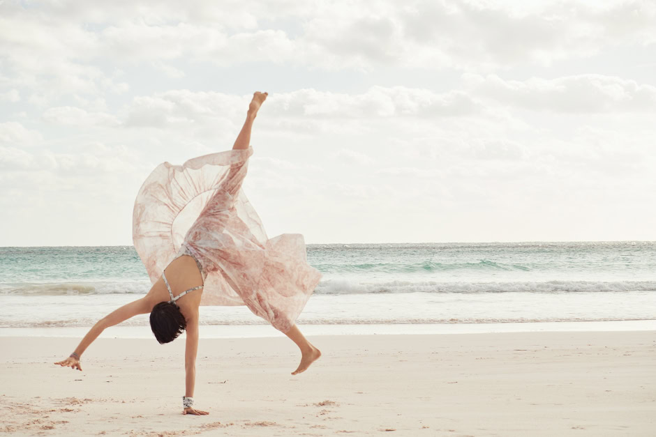 Elegant woman performing a handstand on the beach, wearing a flowing pink dress, with ocean waves and a cloudy sky in the background, showcasing artistic beach photography and lifestyle imagery.
