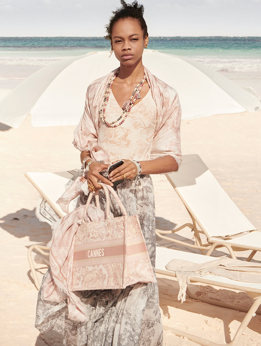 Elegant beach fashion portrait featuring a woman in stylish attire with accessories, holding a Cannes-themed tote bag, on a sandy beach with lounge chairs and ocean background.