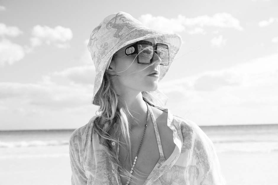 Elegant black and white portrait of a stylish woman wearing oversized sunglasses and a sun hat at the beach, showcasing fashion photography and seaside lifestyle.