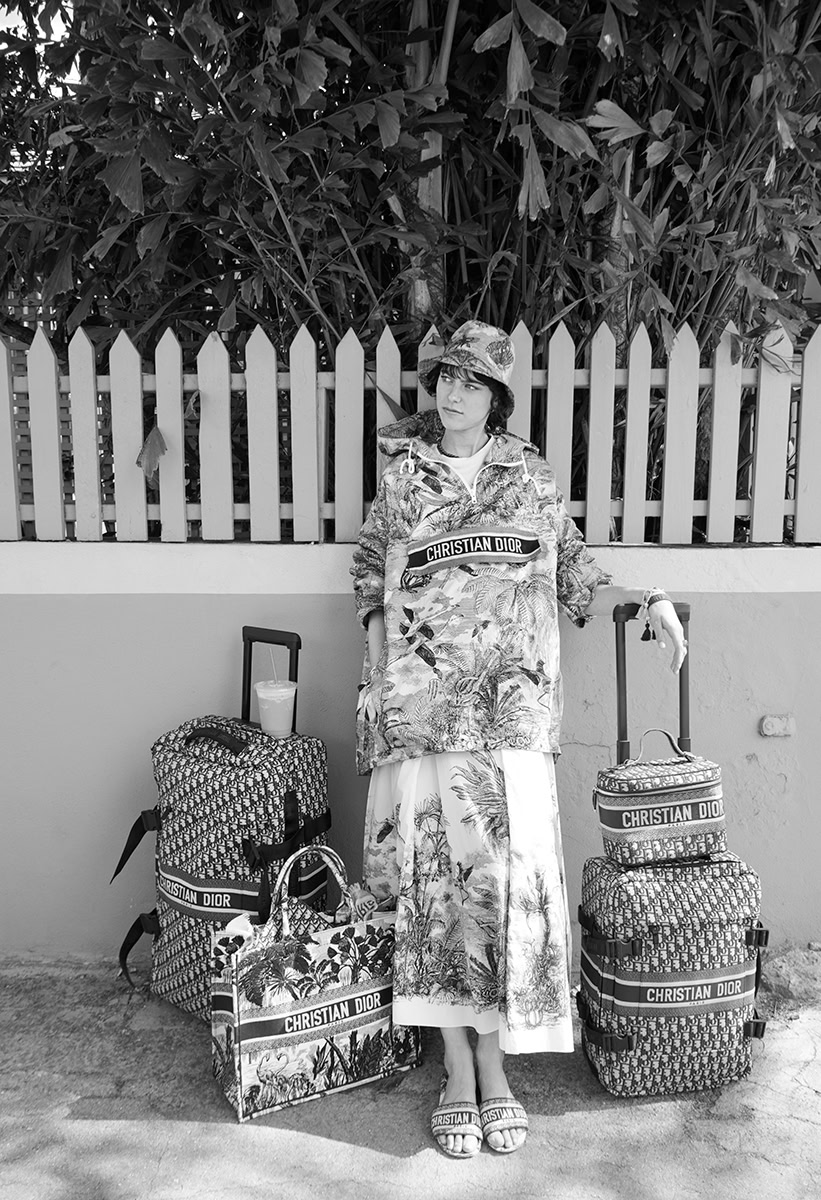 Travel photographer standing outdoors with Christian Dior luggage and tote bags, dressed in tropical print outfit, near a wooden fence and lush greenery.