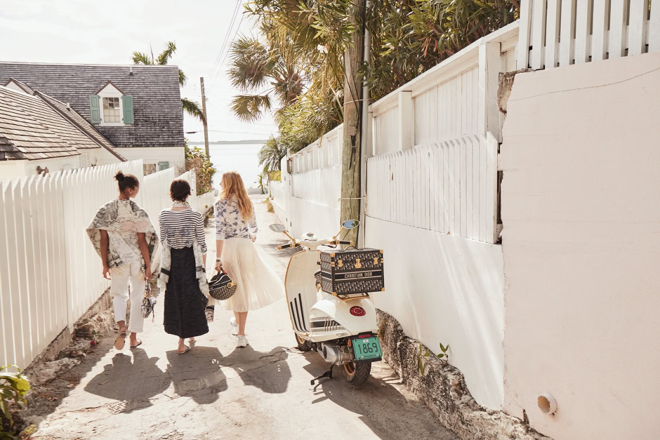 Elegant women walking along a sunlit alleyway with a vintage scooter, featuring tropical surroundings and white fences, capturing a stylish and relaxed summer mood.