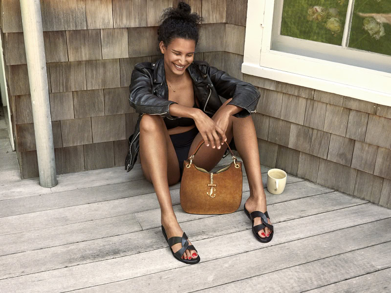 Bright smiling woman sitting outdoors, wearing a black leather jacket and sandals, with a tan handbag and cup, showcasing lifestyle photography by Pamela Hanson.