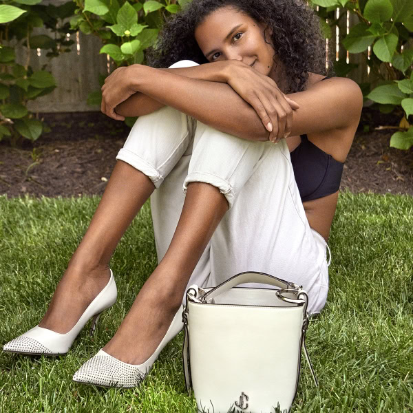 Elegant woman sitting on grass with white heels, black top, cream pants, and stylish white handbag, photo by Pamela Hanson Photography, showcasing fashion, beauty, and lifestyle photography.