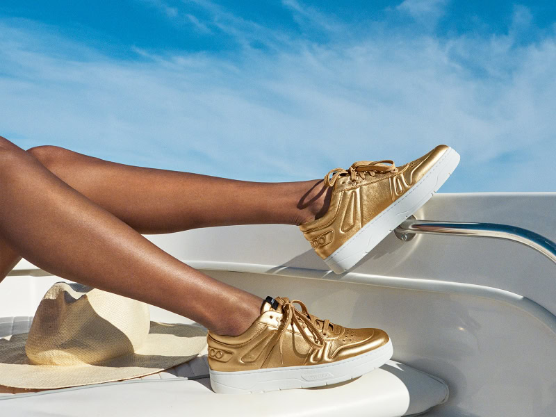 Luxurious gold sneakers on tanned legs relaxing on a white lounge chair under a blue sky, showcasing high-end fashion photography for lifestyle and footwear brands.