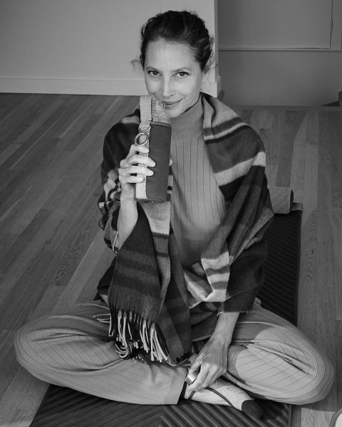 Relaxed woman sitting cross-legged on wooden floor, drinking from water bottle, wearing striped scarf and pinstriped trousers, in candid black and white street photography style.
