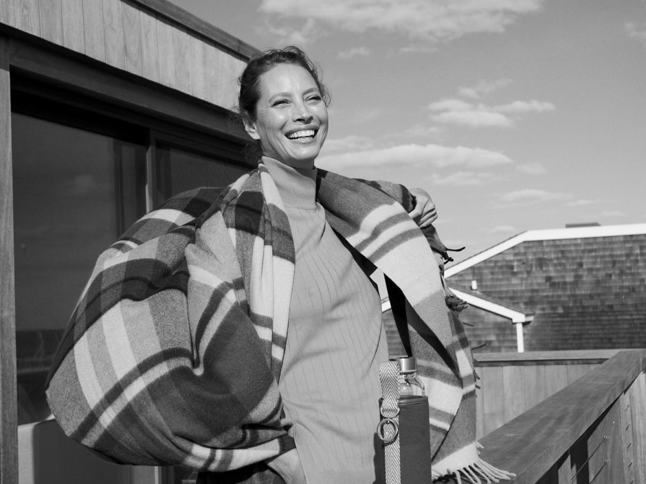 Elegant woman smiling outdoors, wearing a checkered coat and carrying a water bottle, captured in this black-and-white portrait emphasizing fashion and lifestyle photography.
