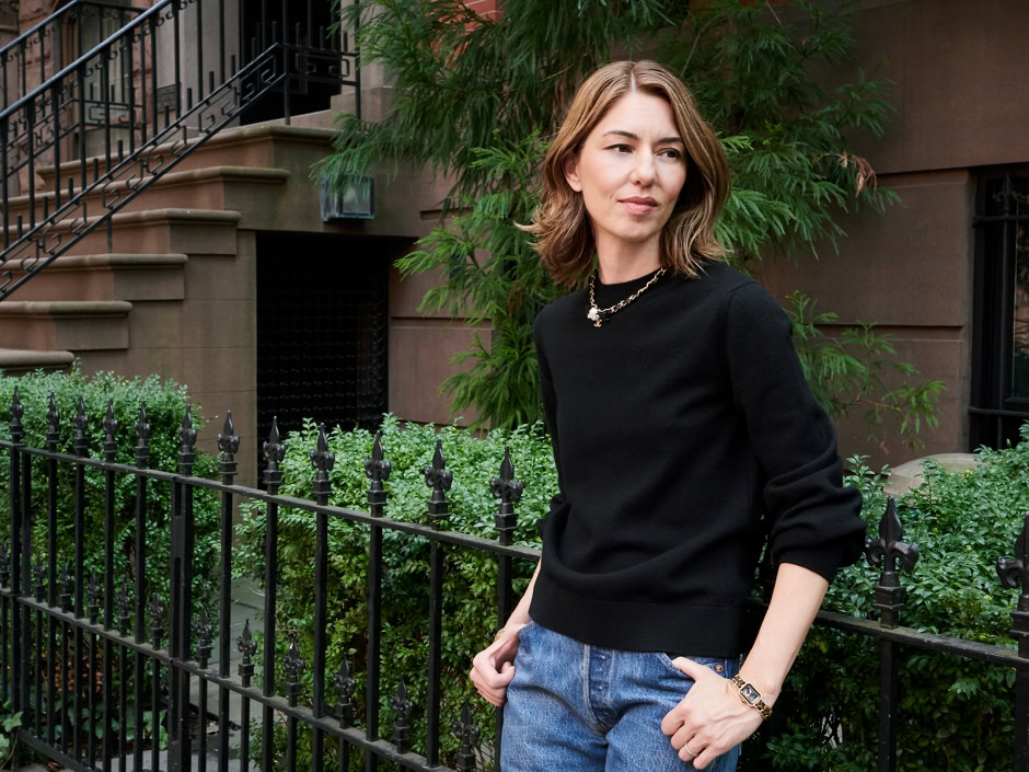 Elegant woman in black sweater and denim jeans posing outdoors in front of green foliage and brownstone building, showcasing contemporary street style in urban environment.