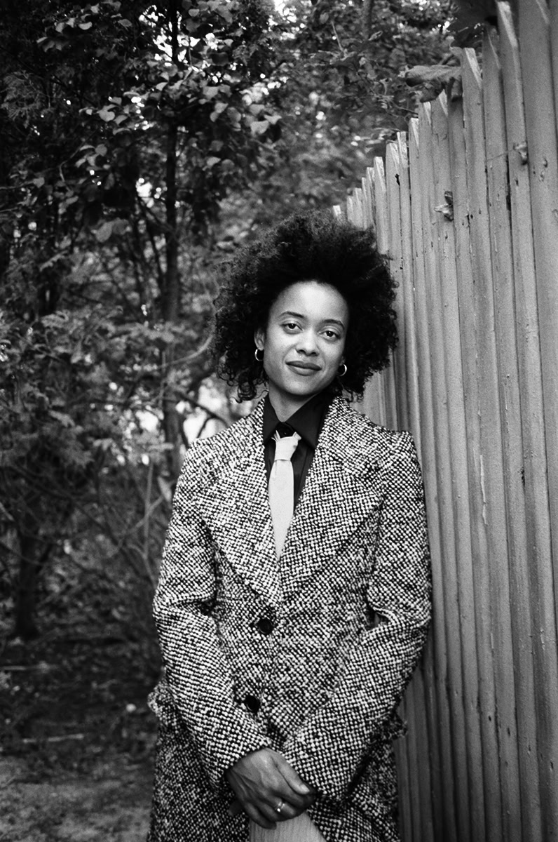 Black and white portrait of a confident woman with natural curly hair, wearing a textured blazer, standing outdoors next to a wooden fence with trees in the background, captured in professional fashion photography style.