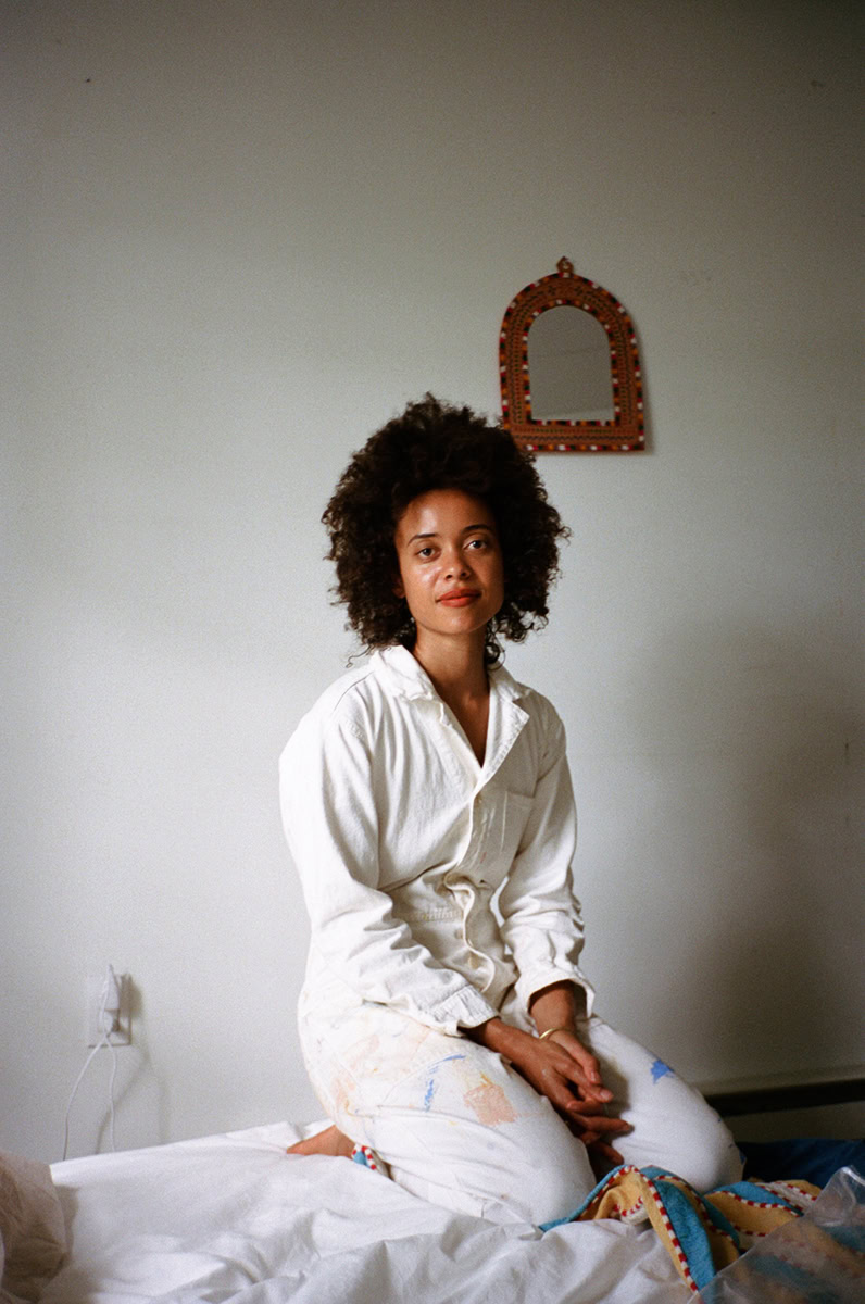 Elegant portrait of a woman with natural curly hair wearing a white shirt, sitting on a bed in a minimally decorated room, showcasing contemporary lifestyle photography by Pamela Hanson.