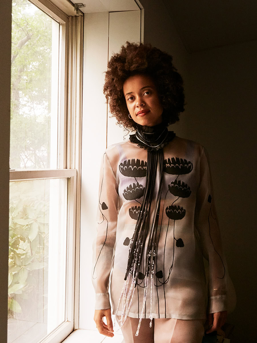 Vibrant fashion photograph of a woman with curly hair wearing a sheer, embroidered blouse with floral motifs, standing by a window with natural light in a modern studio setting.