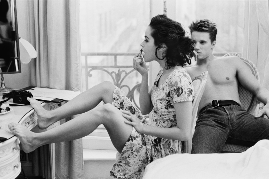 Elegant black and white portrait of a woman applying lipstick while a shirtless young man relaxes in a vintage-style room, showcasing intimate portrait photography by Pamela Hanson.