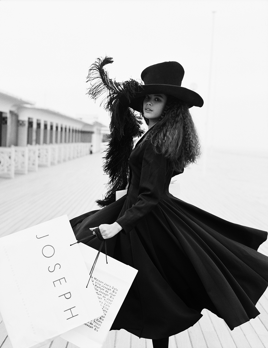 Elegant black-and-white fashion photography featuring a woman in a vintage-inspired outfit with a wide-brimmed hat and feather accents, holding shopping bags on a boardwalk.