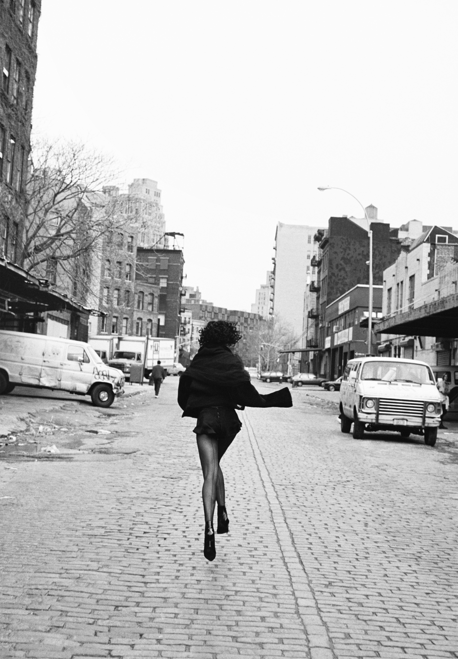 Running woman on cobblestone street in urban cityscape with buildings and parked vehicles, black-and-white fashion photography by Pamela Hanson.