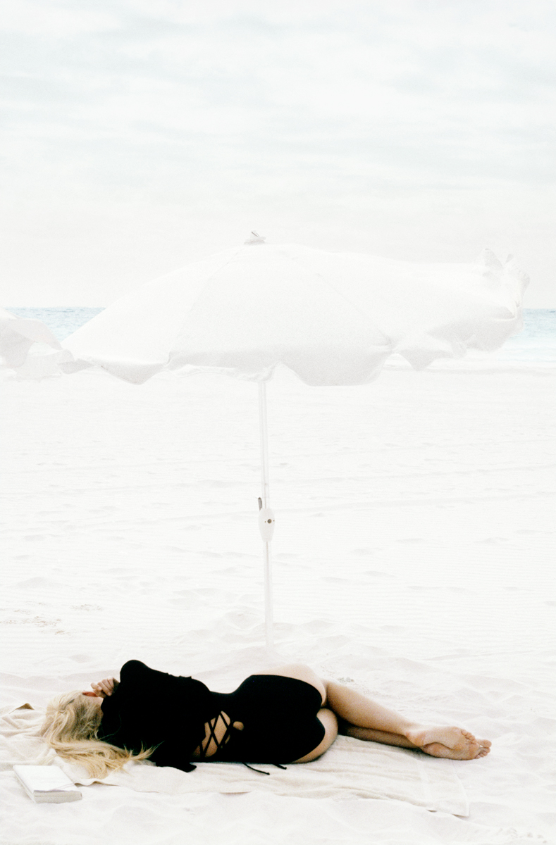 Soft-focus beach photography by Pamela Hanson, featuring a woman under a white parasol on sandy shore. Elegant, artistic portrait emphasizing beach lifestyle, fashion, and relaxation.