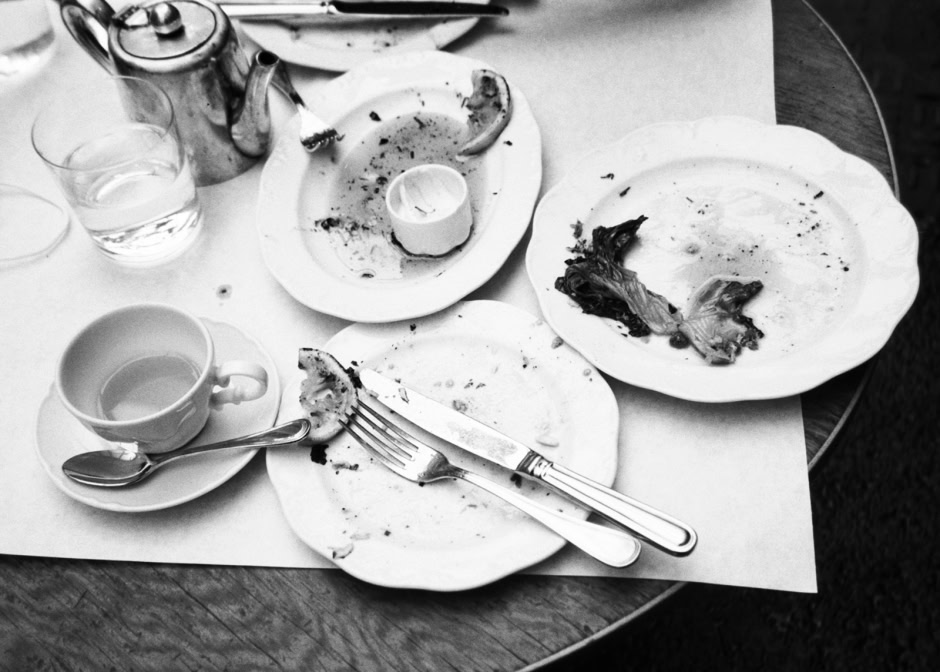Discarded plates with leftover food, knife, fork, cup, and glasses on a table, capturing a candid moment of a finished meal in black and white.