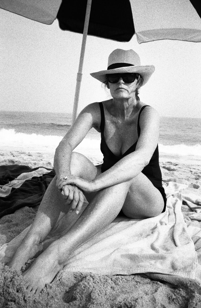 Elegant woman in a black swimsuit with wide-brimmed hat and sunglasses sitting on the sandy beach under a large umbrella, capturing a timeless fashion photography shot.