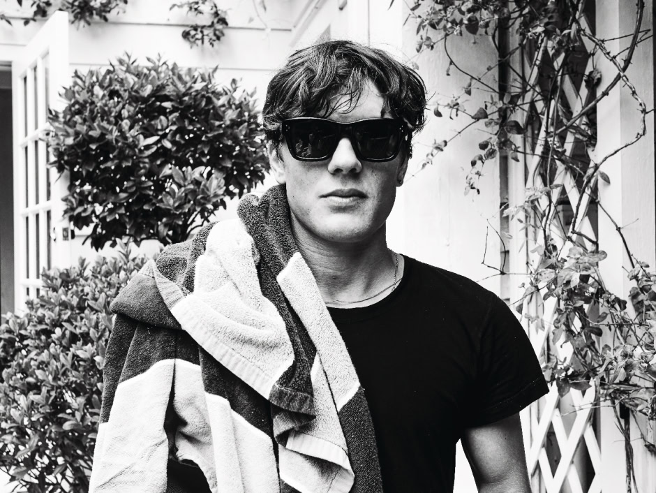 Stylish young man wearing sunglasses with a towel draped over his shoulder, standing outdoors near plants and a white building, capturing contemporary fashion photography.