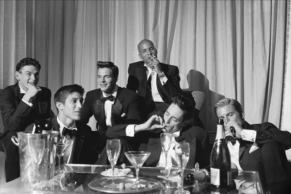 Elegant black and white photograph of a group of young men in tuxedos enjoying a sophisticated party, featuring classic elements like champagne, cigars, and stylish attire, captured by Pamela Hanson Photography.