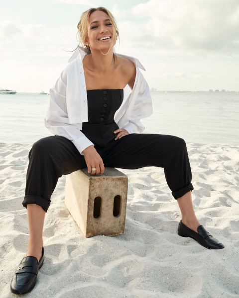 Woman sitting on concrete block on beach, smiling, fashion photography, sunny day, waterfront backdrop, casual yet chic style, professional portrait, Pamela Hanson Photography, beach lifestyle photography.