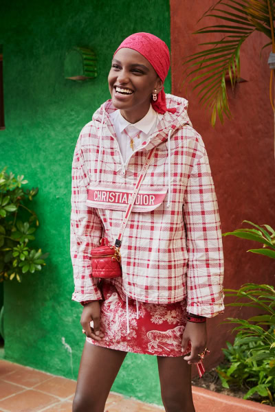Vibrant fashion portrait of a joyful young woman wearing a Dior hoodie, pink headscarf, and floral skirt, standing outdoors with lush green plants and colorful walls.
