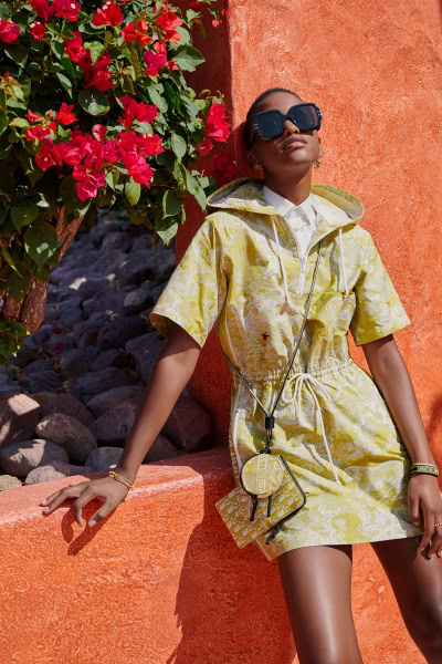 Bold woman wearing oversized sunglasses and a stylish yellow patterned dress, sitting against a vibrant orange wall with pink bougainvillaea, showcasing outdoor fashion photography by Pamela Hanson.