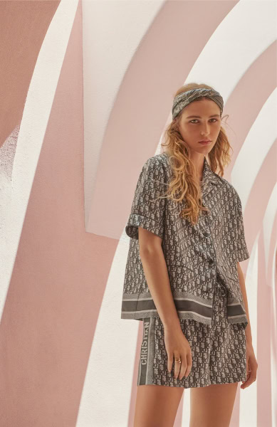 Fashion model wearing Dior pattern matching shirt and shorts, styled with a headband, against a pastel pink architectural background, showcasing luxury fashion photography.