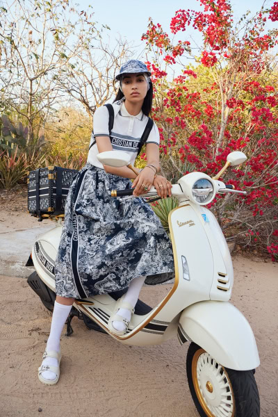 Vespa scooter with a female model in fashionable attire, outdoor setting with vibrant red flowers, fashion photography, luxury lifestyle, Pamela Hanson photography, editorial fashion shoot, street style, high-end fashion image.