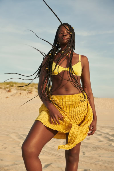 Vibrant woman in yellow swimsuit and skirt posing on beach with breezy hair, natural sunlight, and sandy dunes in the background, showcasing stunning beach fashion photography by Pamela Hanson.