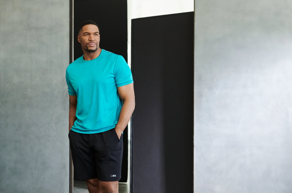 Vibrant portrait of a confident man wearing a turquoise athletic t-shirt and black shorts with hands in pockets, standing indoors near a modern black and gray wall, showcasing contemporary athletic fashion and lifestyle photography.