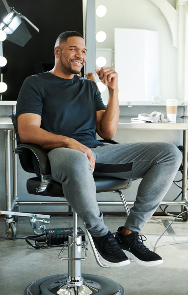 Happy man sitting in makeup chair at studio, professional photo shoot, confident smile, modern photography setting, Pamela Hanson Photography, beauty lighting, male model, portrait photography, creative studio shot, high-quality image, fashion and lifestyle photography, digital photography, editorial portrait.