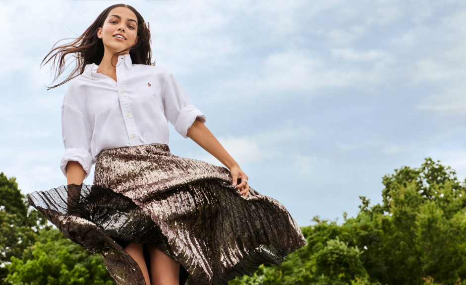 Elegant young woman outdoors, wearing a stylish white shirt and metallic pleated skirt, capturing a fashionable and dynamic portrait with lush greenery and blue sky background.