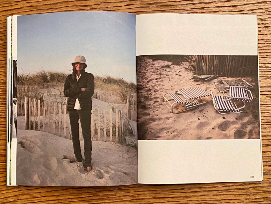 Woman standing on sandy beach with dunes and wooden fence, wearing sun hat and dark clothing, capturing a coastal scenery; relaxed beach vibe.